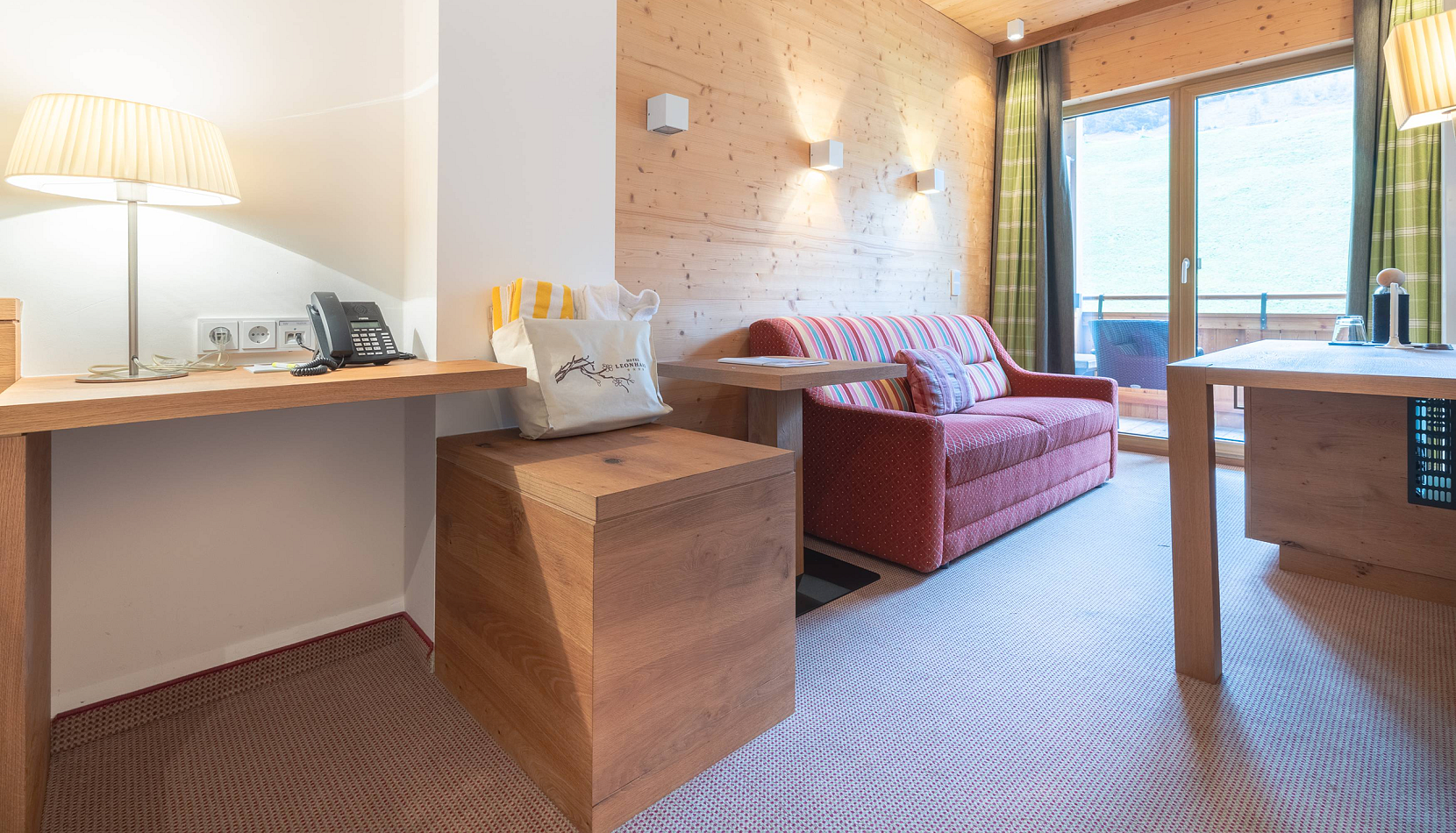 Ein gemütlich eingerichtetes Zimmer in einem alpinen Stil. Das Zimmer hat Holzwände und einen Holztisch mit einer Lampe darauf. Es gibt ein rotes Sofa und einen Balkon mit Blick ins Freie. Die Einrichtung wirkt modern und einladend.