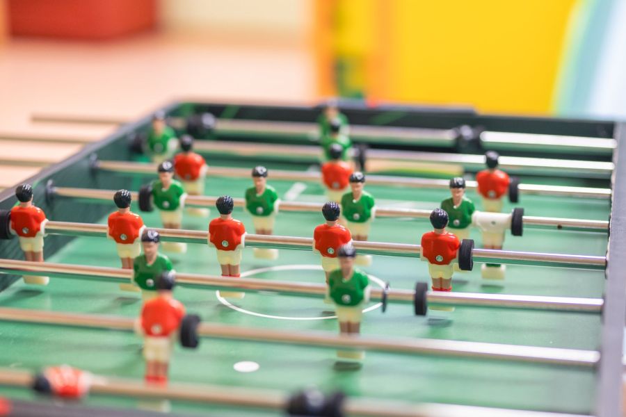 Das Bild zeigt einen Tischkicker, auch bekannt als Tischfußballspiel, mit Figuren in grünen und roten Trikots auf einem grünen Spielfeld. Mehrere Spielfiguren sind an Metallstangen befestigt, die über das Feld verlaufen.