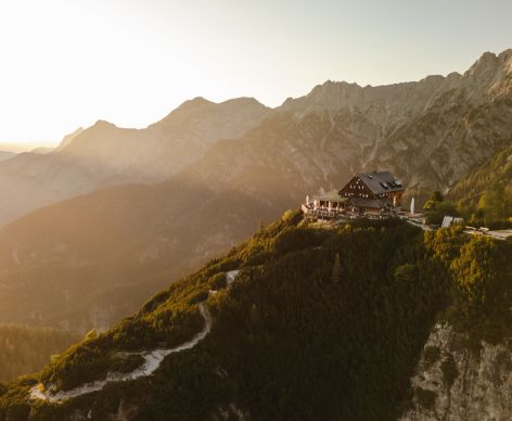 A mountain chalet located on a rocky hillside, surrounded by lush green forests and rugged mountains in the background. The setting sun casts a warm golden light over the landscape, creating a serene and picturesque scene.