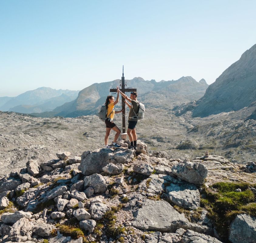 A man and a woman reaching the peak of a rocky mountain, celebrating with a high-five next to a cross. They are wearing backpacks and hiking gear, standing with a scenic view of mountains in the background under a clear blue sky.