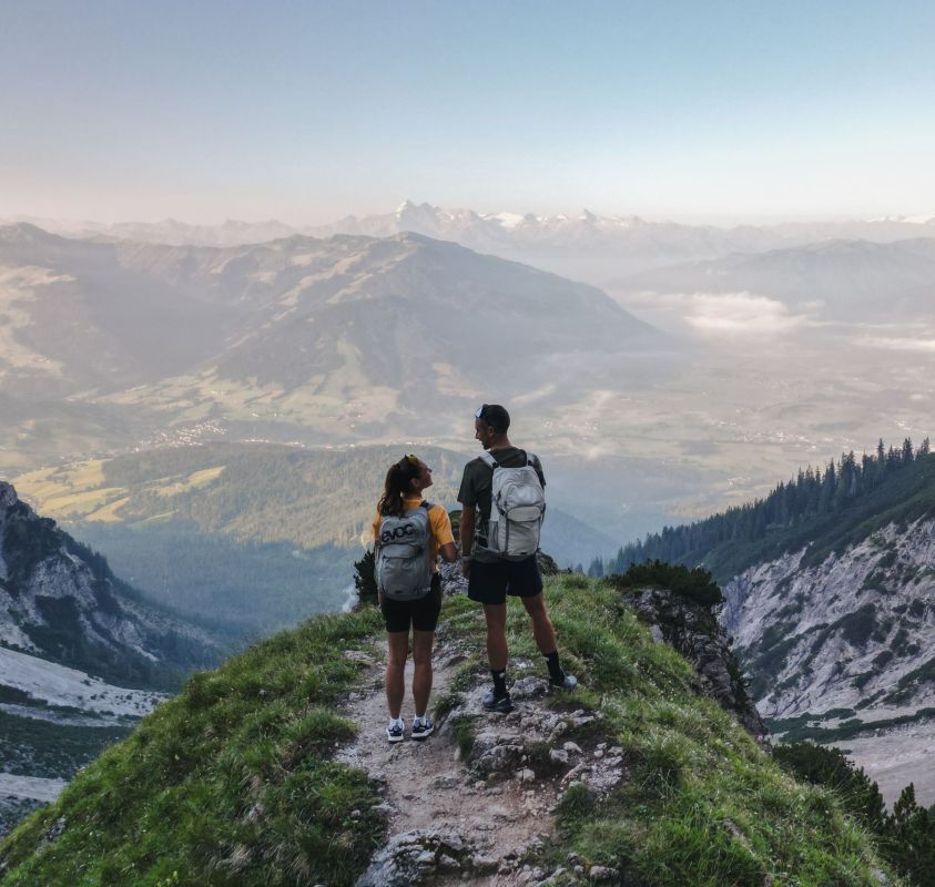 Two hikers with backpacks are standing on a narrow grassy path on a mountainside, overlooking a vast valley with distant peaks under a clear blue sky. The landscape features rocky slopes and a scenic view of nature.