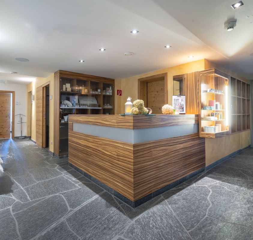A modern reception area in a hotel or spa. The counter is made of wood with a sleek design, and there are shelves with various products displayed. The floor is stone-tiled, and there is modern lighting installed in the ceiling.