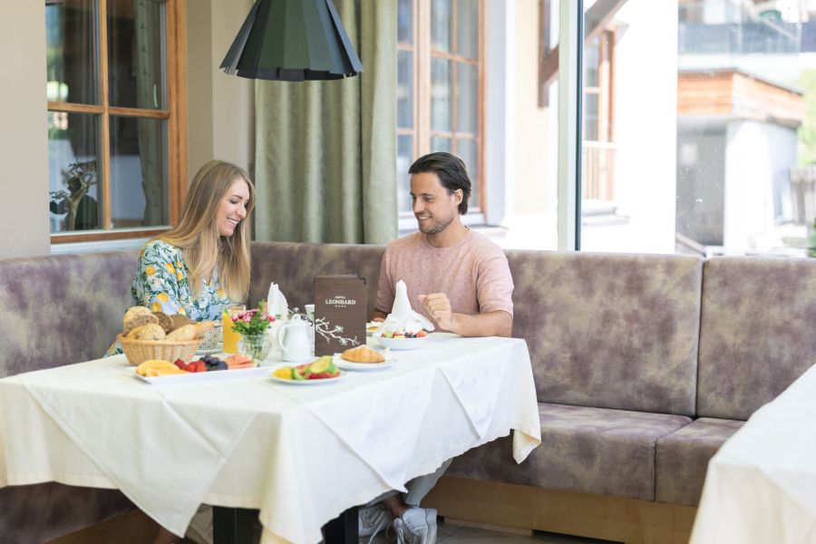 Ein Mann und eine Frau sitzen an einem Tisch in einem Restaurant oder Café. Auf dem Tisch befinden sich Frühstücksgerichte, darunter Croissants, Brot und Obst. Die Umgebung wirkt freundlich und hell mit großen Fenstern im Hintergrund.