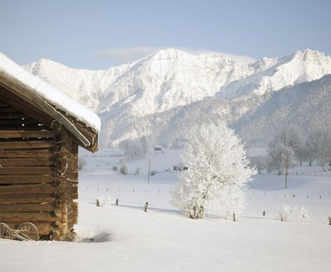 Eine verschneite Landschaft mit einem rustikalen Holzhaus auf der linken Seite, schneebedeckten Bäumen und Bergen im Hintergrund. Der Himmel ist klar und es liegt viel Schnee auf dem Boden.