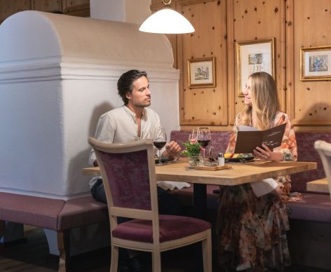 Ein Mann und eine Frau sitzen in einem gemütlichen, traditionell eingerichteten Restaurant an einem Holztisch. Der Tisch ist mit verschiedenen Gerichten und Getränken gedeckt. Die Frau hält ein Menü in der Hand und sie unterhalten sich. Im Hintergrund ist eine Holzvertäfelung zu sehen, die dem Raum eine warme Atmosphäre verleiht.