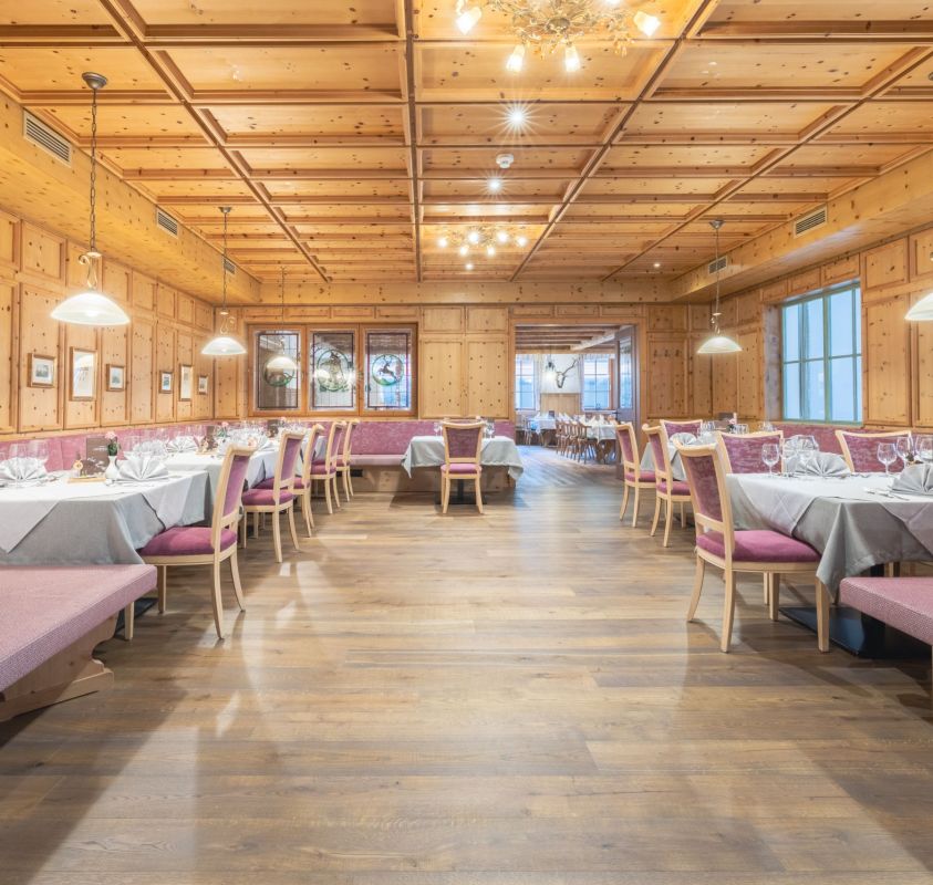Das Bild zeigt ein elegantes Restaurant mit Holzwänden und -decke. Der Raum ist großzügig gestaltet, mit mehreren Tischen und Stühlen, die mit weißen Tischdecken und feinem Geschirr gedeckt sind. An den Seiten befinden sich gepolsterte Sitzbänke. Die Beleuchtung wird durch mehrere hängende Lampen sowie einen zentralen Kronleuchter bereitgestellt. Der Boden besteht aus Holzplanken, und an den Wänden hängen Bilder.