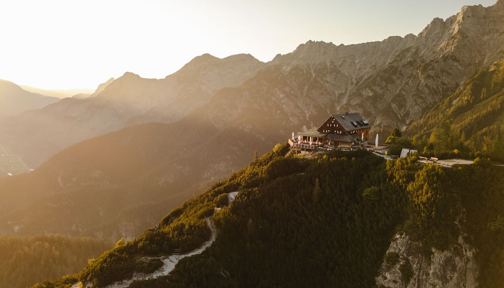 Ein Bergchalet auf einem Bergrücken bei Sonnenuntergang, umgeben von imposanten Berggipfeln und einem Tal.