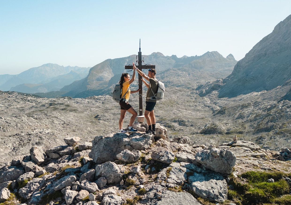A man and a woman reaching the peak of a rocky mountain, celebrating with a high-five next to a cross. They are wearing backpacks and hiking gear, standing with a scenic view of mountains in the background under a clear blue sky.