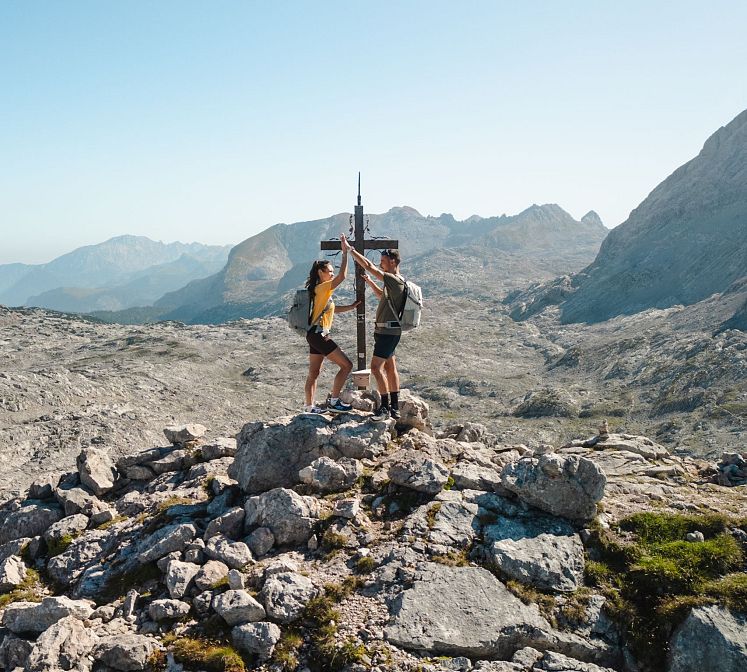 A man and a woman reaching the peak of a rocky mountain, celebrating with a high-five next to a cross. They are wearing backpacks and hiking gear, standing with a scenic view of mountains in the background under a clear blue sky.